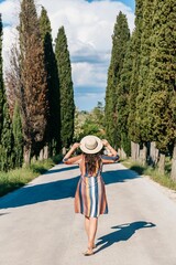 Back view of beautiful woman in dress standing on road surrounded by green trees in Tuscany
