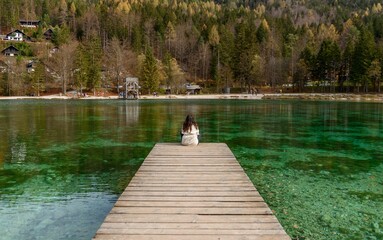 Back shot of a young woman sitting on a wooden pier on the shore of Lake Jasna in Slovenia