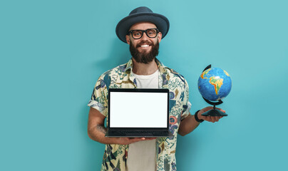 A young guy in a Hawaiian shirt with a beard holds a globe and a laptop in his hand demonstrating a...