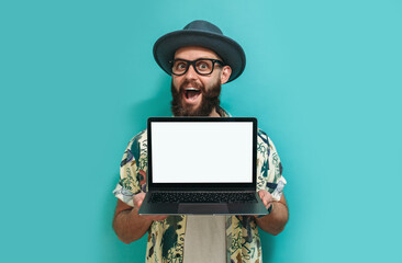 Portrait of a cheerful hipster guy with a beard in a hat and a Hawaiian shirt holding a laptop, showing blank screen with copy space for advertisement and promotional text, demonstrates something. 