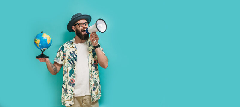 A young guy with a beard in a hat, Hawaiian shirt holds a globe and shouting to the side into a megaphone, isolated on a blue studio background. Travel agency concept
