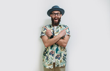 Portrait of positive man in hat and Hawaiian shirt standing with crossed arms and pointing to ad copy space on both sides. Isolated on white studio background