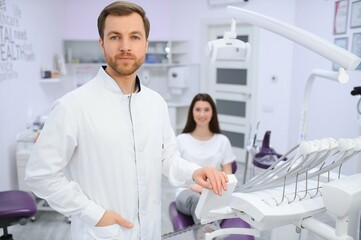 Male dentist in a room with medical equipment and patient on background.