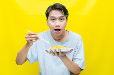 Happy euphoric asian young man in grey tshirt eating instant fried noodle using bamboo chopstick...