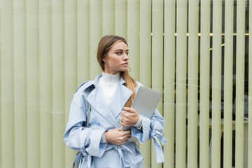 pretty woman in blue trench coat holding laptop while standing near window on street in Vienna.