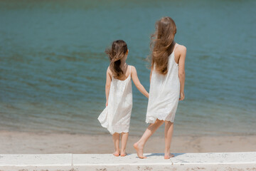 two little girls with long hair in summer dresses are standing on the beach and looking at the sea, rear view