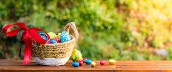 Easter eggs in basket on wooden table banner. Easter season banner with green nature bokeh background. Colorful decorated eggs in wicker basket. Spring season decoration for web banner