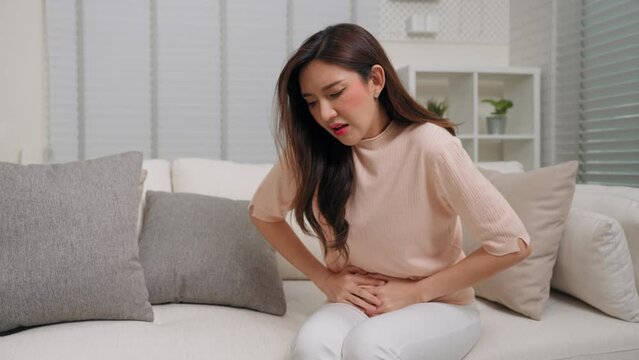Young Asian woman having menstrual pain and sitting on couch in living room at home