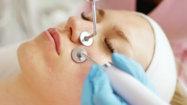 Beautician makes a procedure on the face on the cheekbones and nasolabial folds with microcurrent therapy device. Hardware cosmetology in clinic salon for client.	