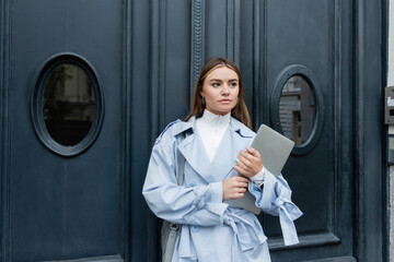 young freelancer in blue trench coat holding laptop while standing on street in Vienna.