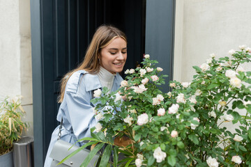 happy young woman in blue trench coat looking at green bush with blooming flowers on street in Vienna.