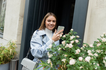 joyful young woman in blue trench coat taking photo of green bush with blooming flowers on street in Vienna.