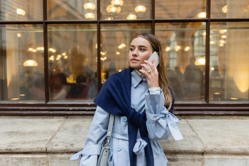 stylish woman with scarf on top of blue trench coat talking on smartphone on street in Vienna.