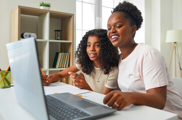 Caring African American mother using laptop to help her daughter with school homework. Smiling mom and preteen girl watching online lesson together sitting at table. Concept for home schooling.