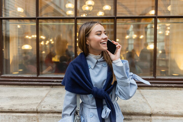 cheerful woman with scarf on blue trench coat talking on smartphone on street in Vienna.