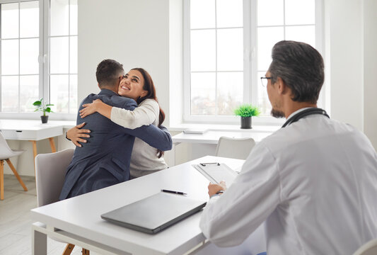 Young Married Couple Are Happy, They Will Soon Become Parents. Good News From Friendly Doctor At Meeting In Office Of Hospital Or Clinic. Joyful Husband And Wife At Pregnancy Planning Consultation.