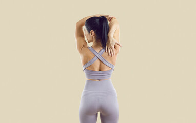 Athletic brunette Caucasian woman stands with back to camera and stretches muscles arms and shoulders before starting sports or fitness workout dressed in gymnastics sportswear posing in beige studio - obrazy, fototapety, plakaty
