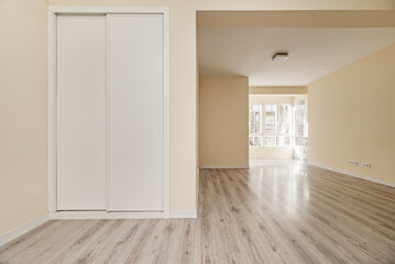 Empty living room with white aluminum and glass windows, light driftwood flooring and a built-in wardrobe with white sliding doors