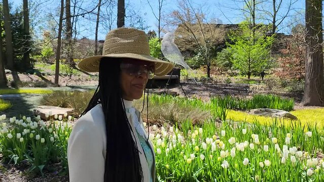 An African American woman wearing a hat and sunglass walking through a gorgeous spring landscape in the garden with lush green trees and plants and colorful flowers at Atlanta Botanical Garden