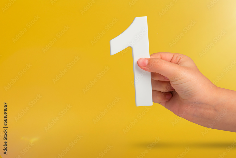 Aufkleber Number one in hand. A hand holds a white number one on a yellow background with copy space. Concept with number one. 1 percent rate, birthday, first or winner.