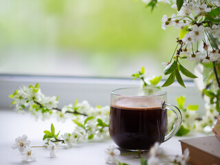 Cup of coffee on the window, green spring background, white cherry blossoms, copy space