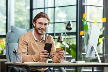 Happy young man businessman received monetary reward, transaction, investment, online shopping. Sitting at the desk in the office, holding a credit card and smiling at the camera.