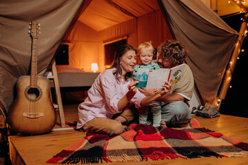 Obraz na płótnie Canvas Happy family with lovely baby playing and read book together in glamping on summer evening. Luxury camping tent for outdoor recreation and recreation. Lifestyle concept