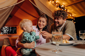 Happy family with lovely baby have dinner and spend time together in glamping on summer evening near cozy bonfire. Luxury camping tent for outdoor recreation and recreation. Lifestyle concept