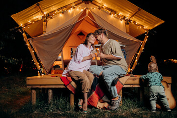 Happy couple relaxing in glamping on summer evening and drinking wine near cozy bonfire. Luxury camping tent for outdoor recreation and recreation. Lifestyle concept