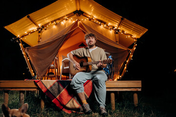 Obraz na płótnie Canvas Handsome young smiling man playing guitar sitting in cozy glamping tent in summer evening bonfire. Luxury camping tent for outdoor holiday and vacation. Lifestyle concept