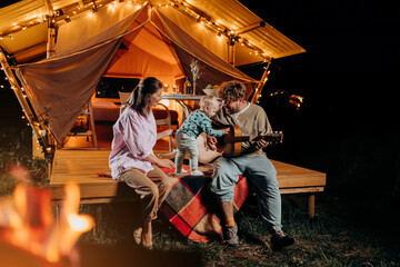 Happy family relaxing and spend time together in glamping on summer evening and playing guitar near cozy bonfire. Luxury camping tent for outdoor recreation and recreation. Lifestyle concept - 581915273