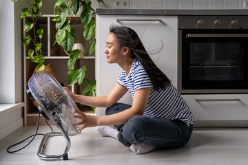 Overheating at home. Happy joyful young Asian woman sitting in front of electric floor fan at home, cooling down, refreshing during hot summer days in apartment without air conditioner in summer