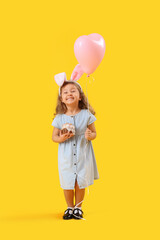 Little girl in bunny ears with Easter cake and balloon on yellow background