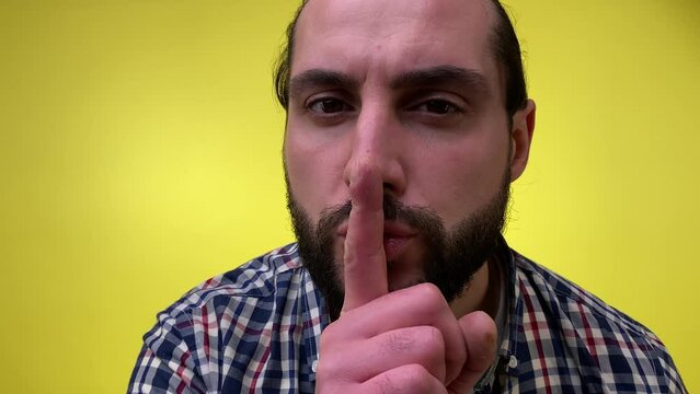 Close-up portrait mysterious bearded man in casual clothing saying hush be quiet with finger on lips shhh gesture isolated on yellow background. Secret middle-aged male person approaching close camera