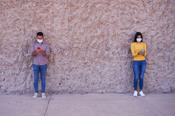 Obraz na płótnie Canvas Young couple wearing masks using mobile phones and maintaining social distance while standing against wall on street