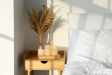 Burning candles, book and vase with pampas grass on table near light wall