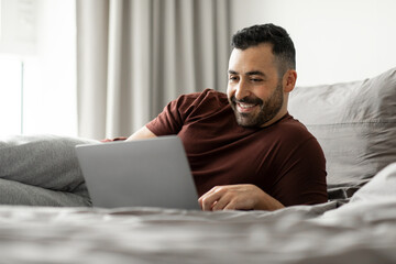 Happy man using laptop lying in bed in bedroom interior, working from home on weekend, browsing internet and typing, free space