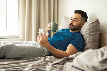 Fototapeta na wymiar European man using cellphone and drinking coffee, holding cup and browsing internet while sitting in bed in modern bedroom indoor, free space