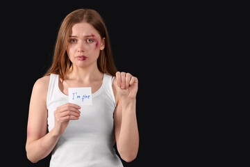 Bruised young woman holding paper with text I'M FINE on dark background. Domestic violence concept