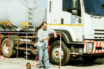 Asian male worker driving truck transport oil business industry in regional gas station checking car time on tablet parking lot to travel delivering liquid oil is on its way to destination station.