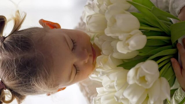 Little girl sitting by window with tulip flowers bouquet. Happy child, indoors. Mother's day, valentine's day or birthday concept.