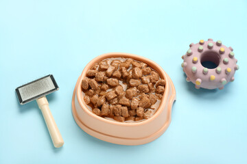Bowl of wet pet food, toy and grooming brush on color background