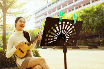 Beautiful asian teacher sits and plays ukulele in front of school building preparing to teach...