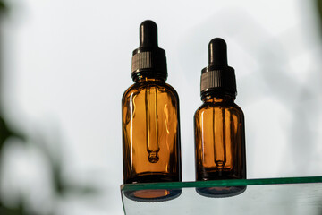 Serum in glass bottle with dropper lid on glass stand. Essential oil for care of women's skin on white background, in rays of sunlight. Space for copying, glare on object.
