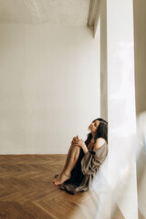 young beautiful woman sits on the floor with her back against the wall. a brunette girl sits on a parquet floor in a room with white walls and dreams about something.