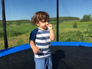 curly cute tender boy smiling child with a finger in his mouth stands on a trampoline