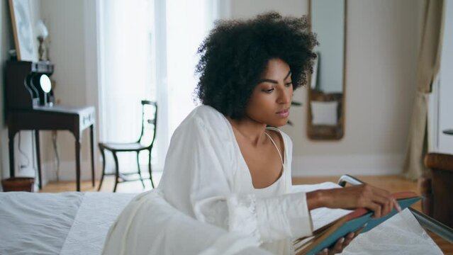 Peaceful girl read novel bed alone. African woman laying bedroom flipping pages