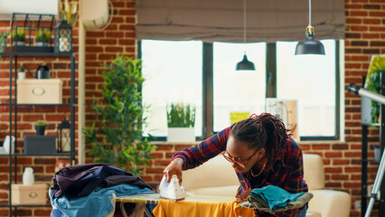 Obraz premium African american woman using steam iron and ironing board, smoothing out creases on clothes. Female person doing household chores for spring cleaning, using laundered clothing.