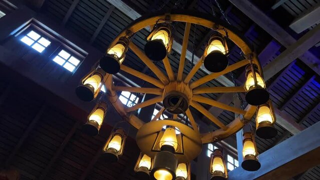 An antique wooden chandelier in a wooden tall building with small windows under the ceiling hangs gloomy and shines with 12 lamps similar to fire. High quality 4k footage
