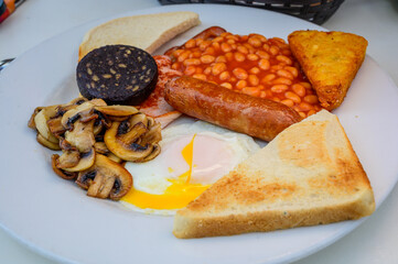 White board with full English breakfast with bacon, fried egg, beans, tomato, roasted sausage, black pudding, scons, hash browns and fried mushrooms
